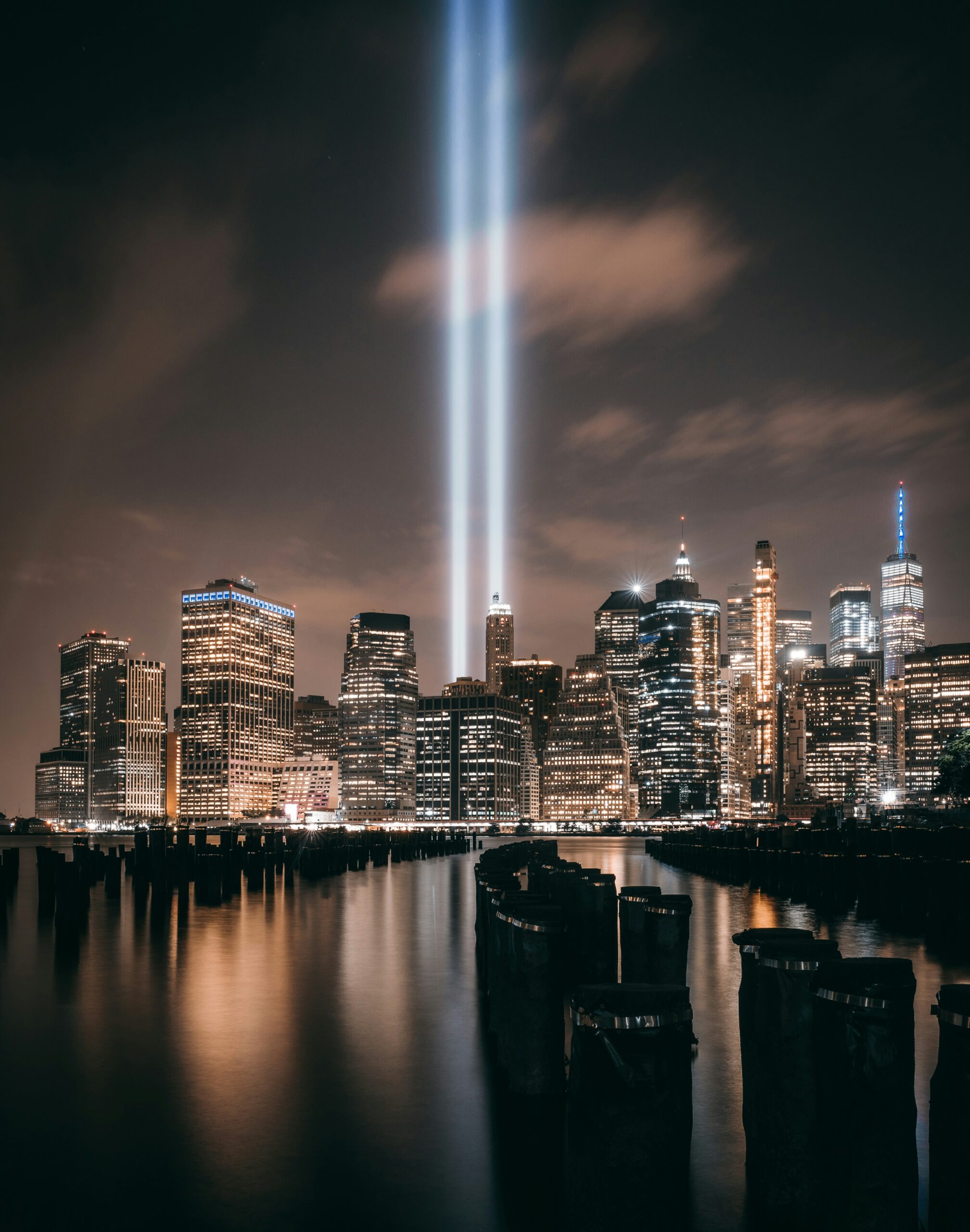 Two beams of light rising into the night sky above the illuminated Manhattan skyline, reflected in the water.