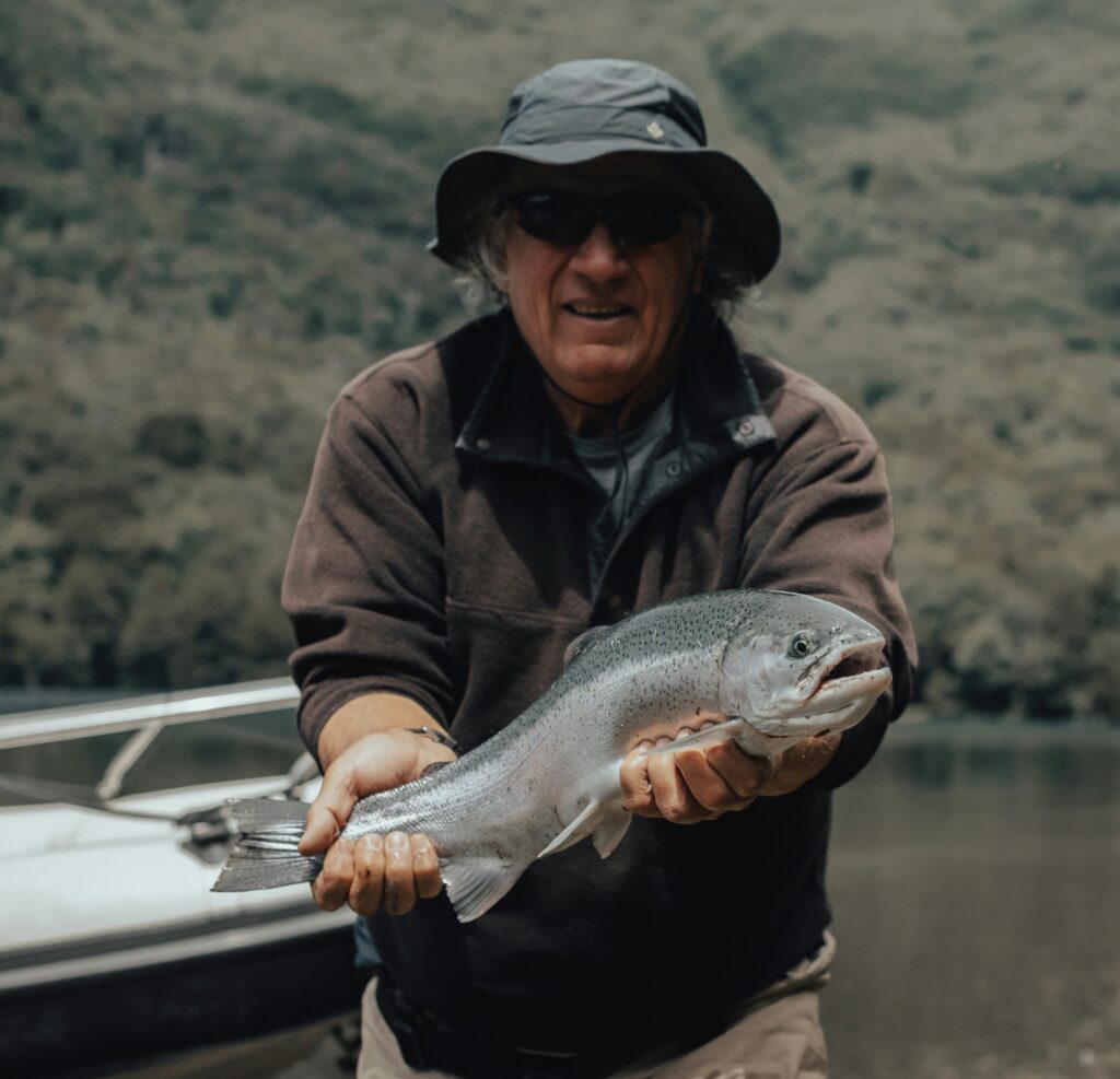 An older man in a bucket hat holding up a freshly caught fish